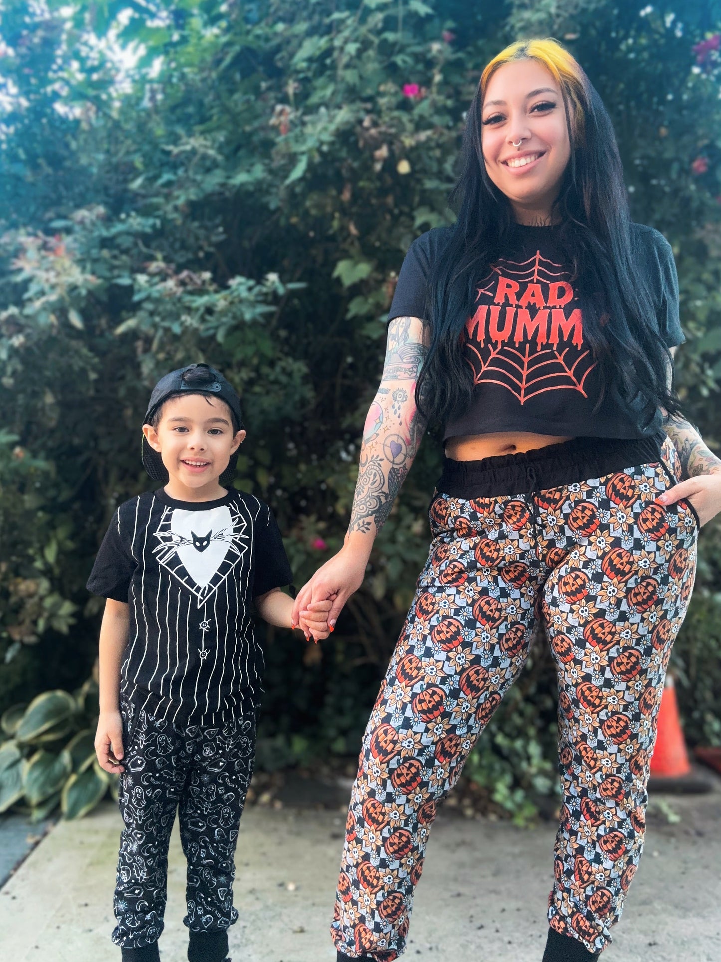 Two people, a woman and a child, standing outdoors holding hands with Halloween-themed outfits.