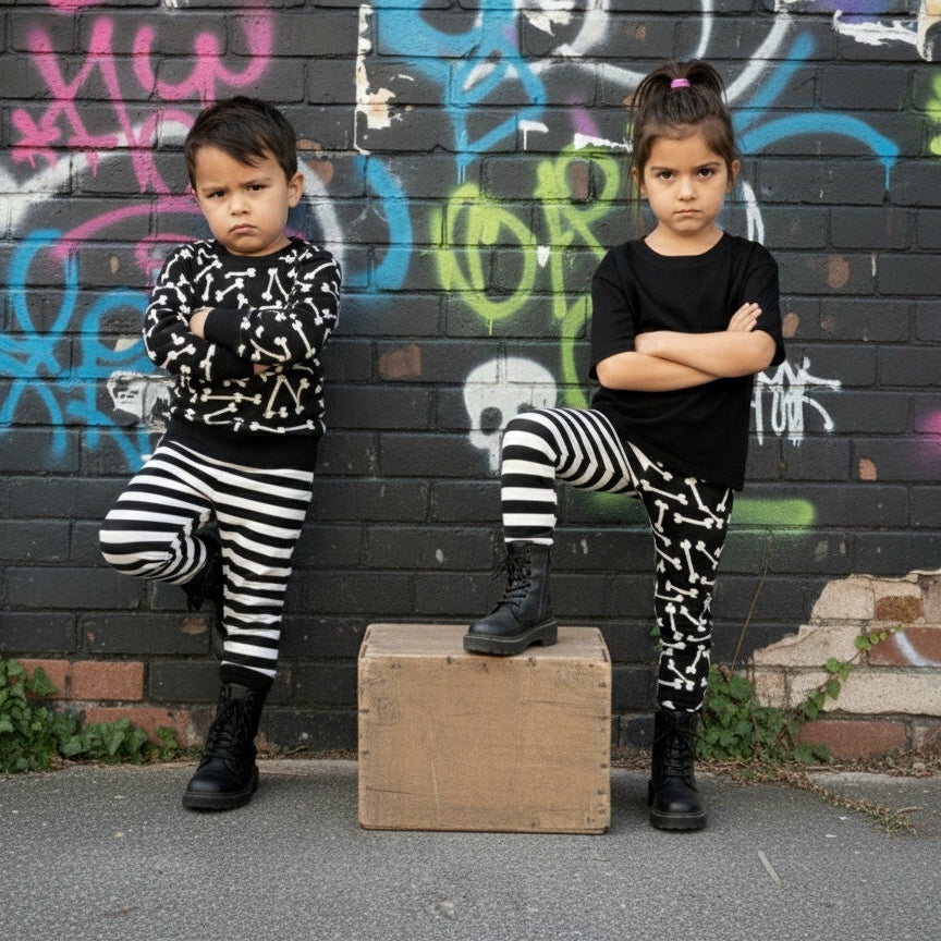 Two children, goth, punk style, in black and white outfits standing against a graffiti-covered wall.
