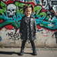 Child wearing a black leather jacket and black dress with big white bones standing in front of a graffiti-covered wall.