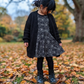 A young girl standing in an autumn park, wearing a black knit cardigan over a black and white spiderweb-patterned dress, black tights, and black boots on a ground covered in fallen leaves.