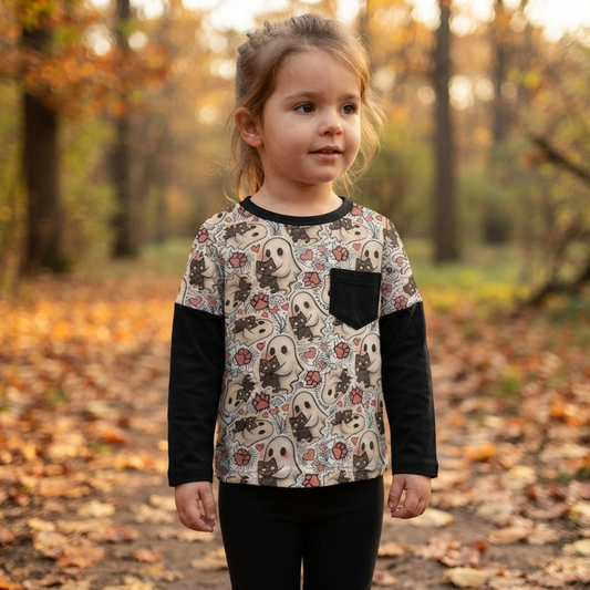 A young girl stands on a leaf-covered forest path wearing a long-sleeve top with black sleeves and a patterned front panel featuring a pocket, photographed outdoors in autumn.