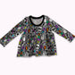 A long-sleeve baby/toddler top with a gray base covered in a colorful Halloween pattern of skulls, ghosts, lightning bolts, and tombstones, featuring a black crew neck.
