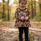 A young child standing in a wooded outdoor setting, wearing a long-sleeve black top with an orange and beige pumpkin pattern, black pants, and black boots.